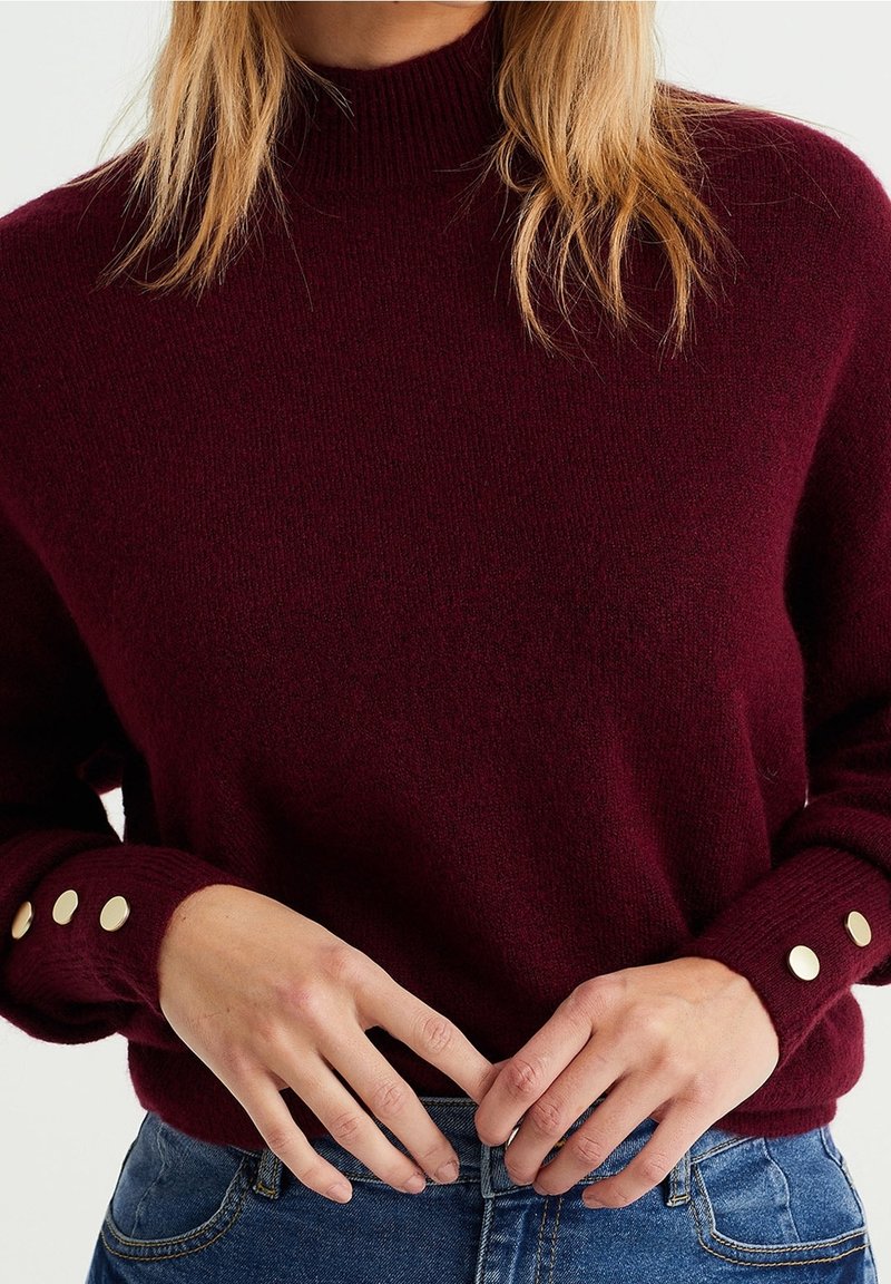 Burgundská pletená svetr s vysokým límcem, který má zlaté knoflíkové detaily na manžetách a jemnou texturu. Spárováno s modrými džíny.