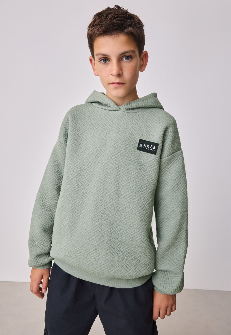 Garçon aux cheveux courts et bruns portant un sweat à capuche vert clair texturé et un pantalon foncé, debout devant un fond clair uni.