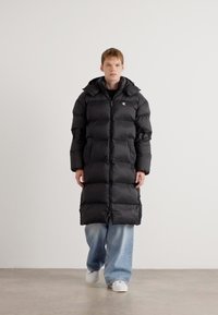 Sort, polstret lang frakke med hætte, med en frontlynlås og elastiske manchetter, båret over en sort top og løse blå jeans. Hvide sneakers.