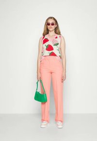 Colete de malha com padrões de frutas vermelhas e rosas, combinado com calças claras rosa. Os acessórios incluem uma bolsa verde de ombro e óculos de sol.