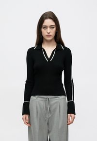 Camisola preta canelada com decote em V e vivo branco ao longo da gola e das mangas, combinada com calças cinza claro de corte largo.