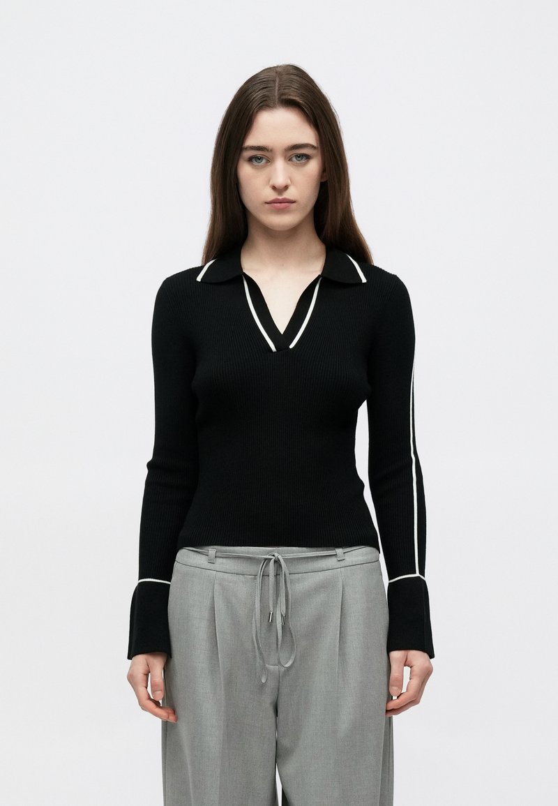 Camisola preta canelada com decote em V e vivo branco ao longo da gola e das mangas, combinada com calças cinza claro de corte largo.