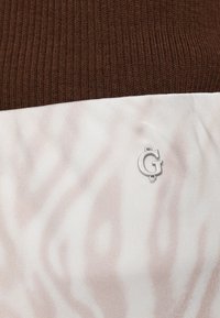 Smeđa pletena tkanina na vrhu prelazi u glatku kremastu tkaninu s diskretnim ružičastim zebrinim prugama. Srebrni logo je smješten u središnjem dijelu donjeg dijela.