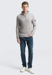 Pull en maille torsadée gris clair avec un col haut, assorti à un jean bleu foncé et des baskets blanches. Comprend des poignets et un ourlet côtelés.