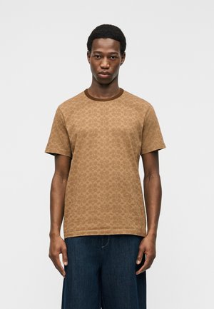 Man wearing a brown patterned short-sleeve t-shirt and dark blue pants, standing against a plain light background.