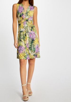 Femme portant une robe sans manches longueur genou avec un motif de feuilles tropicales jaunes, vertes et violettes, associée à des sandales compensées beige à bride cheville.