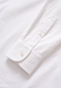 Polsino di camicia bianca in cotone con bordo arrotondato, dotato di un solo bottone e una texture liscia. Il bottone è in plastica.