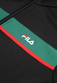 Chaqueta negra con rayas horizontales verdes y rojas. Incluye un logo blanco de FILA en la zona del pecho. El material parece texturizado y suave.