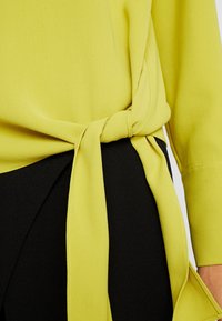 Keltainen, kevyt paita, jossa on sidottu solmukohta vyötäröllä, yhdistetty mustiin kangasvaatteisiin, jotka esittelevät sileää tekstuuria ja yksinkertaista muotoilua.