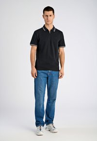 En ung man står vänd framåt iförd en svart pikétröja, blå jeans och vita sneakers mot en ljus, enfärgad bakgrund.