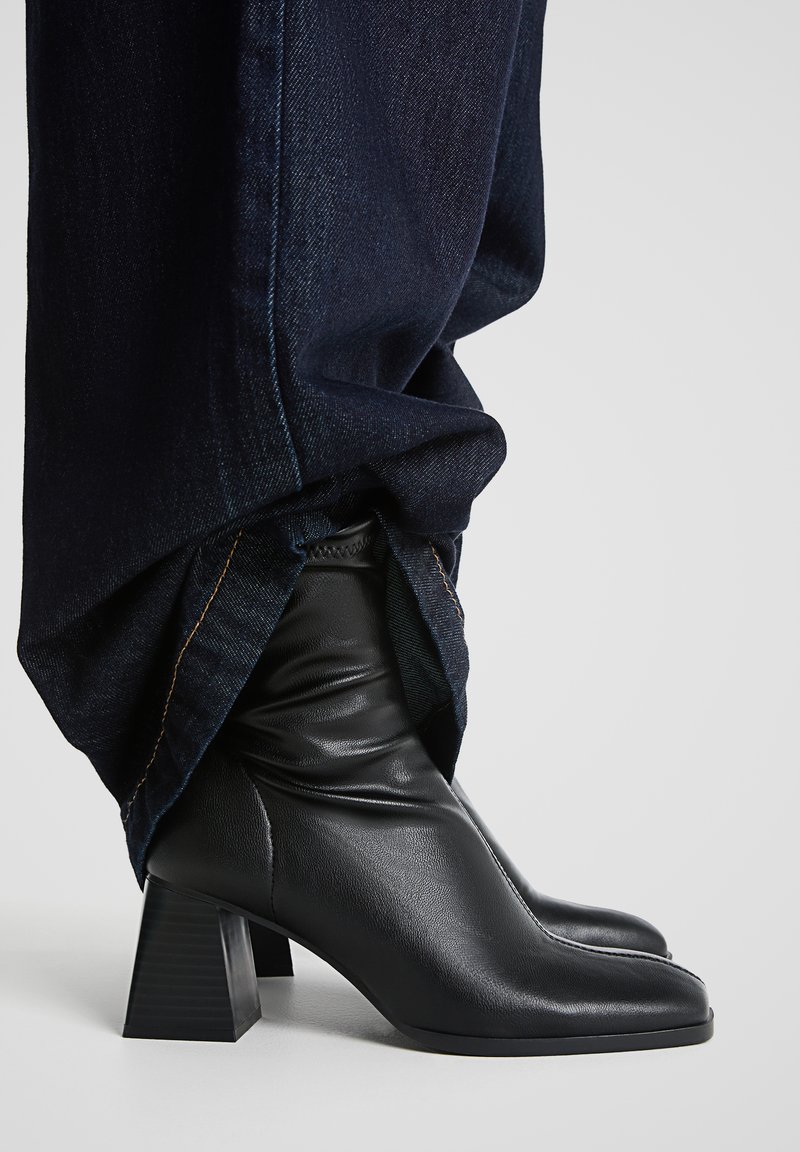 Schwarze Lederstiefeletten mit Blockabsatz, spitzer Zehenform und gerafftem Schaft. Getragen mit dunklen Jeans, die die Stiefel teilweise verdecken.