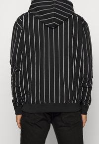 Mies, joka pukeutuu mustaan huppariin, jossa on valkoiset pystysuorat raidat, nähtynä selkäpuolelta, yhdistettyinä mustiin housuihin.