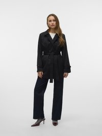 Manteau noir avec des boutons croisés, taille ceinturée et manches longues. Associé à un pantalon en denim large de couleur foncée et des talons bordeaux.
