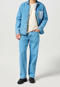 Giacca in denim azzurro chiaro con tasca a toppa, abbinata a pantaloni azzurro chiaro. T-shirt bianca sotto e calzature nere.