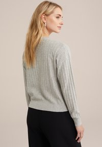 Pilkas raukšlėtas megztinis su ilgomis rankovėmis, prigludusia apačia ir minkšta tekstūra, derinamas su juodomis kelnėmis. Minimalistinis dizainas be matomų logotipų.
