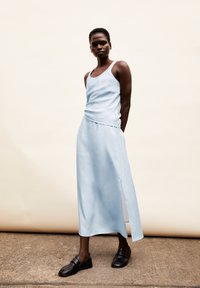 Lichtblauwe, mouwloze jurk met een ronde hals, gedrapeerde stof en een zijsplit, gecombineerd met zwarte instap schoenen op een betonnen oppervlak.