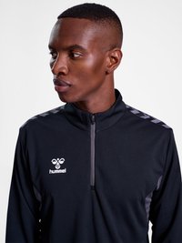 Mężczyzna z krótkimi włosami, ubrany w czarną sportową kurtkę Hummel na zamek błyskawiczny z wzorzystymi paskami na ramionach, patrzy w lewo.