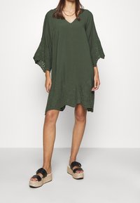 Olivgrünes Kleid mit V-Ausschnitt und weiten, spitzenbesetzten Ärmeln. Dekorativer Laser-Cut-Saum. Paar schwarze Plateausandalen mit Korksohle.