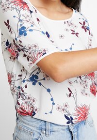 Blomstret bluse med hvid bund, korte ærmer og løs pasform. Designet inkluderer røde, blå og lyserøde blomster med grønne blade.