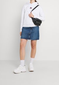 Sudadera blanca con bolsillo frontal, combinada con una falda de mezclilla azul. Bolsa cruzada negra, zapatillas blancas y calcetines tobilleros blancos.