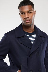 Granatowy wełniany płaszcz peacoat z szerokimi klapami, podwójnymi guzikami i bocznymi kieszeniami; noszony na paskówce. Gładka faktura, zorganizowany krój.