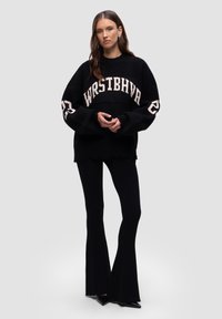 Schwarzer, übergroßer Strickpullover mit dem Schriftzug "WRSTBHVA" in Creme, weiten Ärmeln und rohem Saum; kombiniert mit schwarzen, gerippten Schlaghosen.