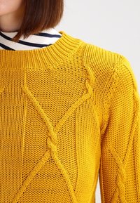 Gul stickad tröja med en strukturerad kabelstickningsmönster, ribbad krage och avslappnad passform. Bakgrunden är helt vit.