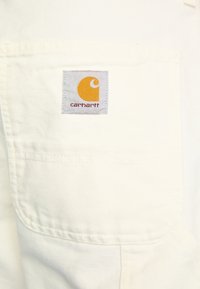 Hvidt stof med en tekstureret overflade, der har en firkantet lap mærket "carhartt" med et gult logo. Enkelts design uden yderligere detaljer.
