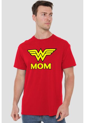 Mann trägt rotes T-Shirt mit gelbem Wonder Woman-Logo und dem Wort "MOM," und steht vor einem einfarbigen hellgrauen Hintergrund, schaut nach rechts.