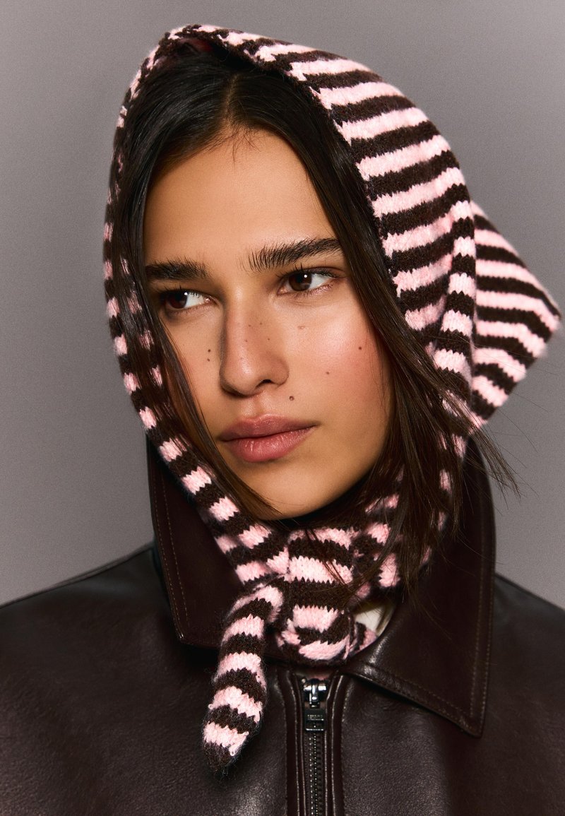 Jeune personne aux longs cheveux bruns portant un foulard en tricot rayé rose et marron ainsi qu'une veste en cuir marron foncé avec une fermeture éclair.