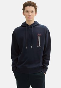 Granatowy hoodie wykonany z materiału o gładkiej fakturze. Posiada grafię z przodu w czerwono-białym tekście, kieszeń kangurką oraz sznurki do regulacji.