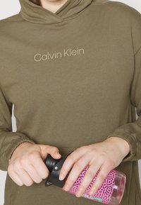 Persona con una sudadera oliva de Calvin Klein abriendo una botella de agua transparente que tiene una funda de patrón púrpura y negro.
