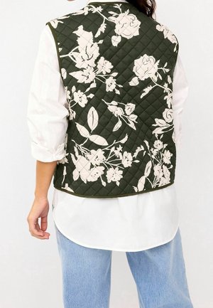 Chaleco acolchado en verde oscuro con ligeros patrones florales. Presenta un dobladillo redondeado, diseño sin cuello y se combina con una camisa blanca y jeans azules.