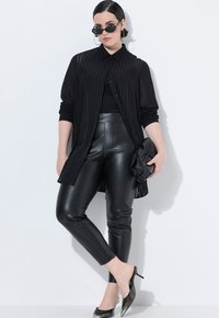 Zwarte geplooide blouse met knopen, strakke zwarte leren broek, zwarte hoge hakken en stijlvolle zonnebril, staand tegen een witte achtergrond.