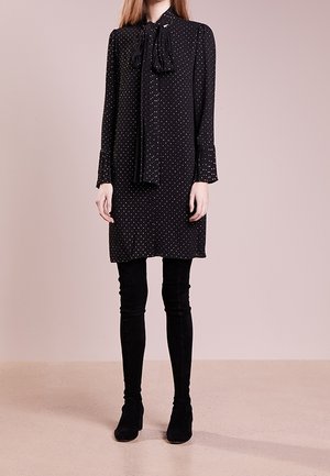 Nainen, jolla on musta pitkähihaisen mekon valkoisilla pilkuilla, kauluksessa solmittu rusetti, mustat legginssit ja mustat nilkkurit.