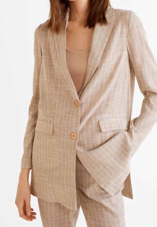 Femme portant un blazer beige à fines rayures avec deux boutons, des poches à rabat et un pantalon assorti, posant avec une main dans la poche.