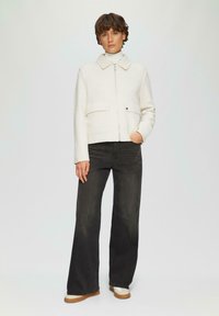 Off-white cropped jack met een kraag, een ritssluiting aan de voorkant en twee borstzakken. Gecombineerd met donkere wijdvallende jeans en beige schoenen. Gladde textuur.