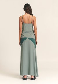 Maxi šaty z tlmeného zeleného materiálu, s tenkými ramienkami, priliehavým živôtikom a kontrastným tmavším zeleným trojuholníkovým panelom na okraji.