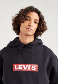 Čierna mikina s kapucňou a sťahovacou šnúrkou, s veľkým červeným logom "LEVI'S" na hrudníku. Mäkká tkanina a voľný strih.