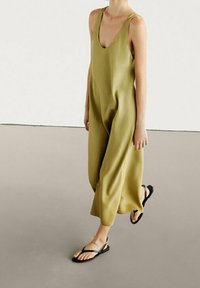Olivengrüner Jumpsuit mit tiefem V-Ausschnitt, weitem Bein und dünnen Trägern. Das Model trägt schwarze flache Sandalen vor einem neutralen Hintergrund.