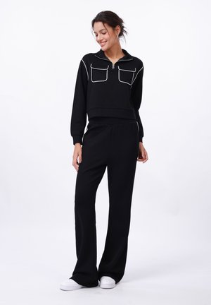 Black sweat top with white piping, zip front, and dual chest pockets paired with loose-fit black pants and white sneakers on a white background.