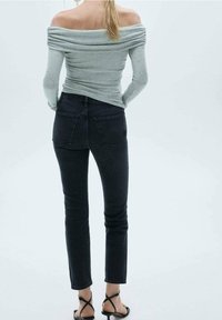 Mujer vestida con un top gris de manga larga y hombros al aire, jeans ajustados negros y sandalias de tacón negro con tiras, de pie frente a un fondo liso.