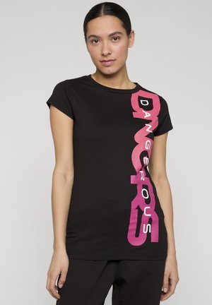 Ung kvinne med mørkt hår iført en svart T-skjorte med fet rosa tekst "DANGEROUS" som går vertikalt på høyre side, stående mot en ensfarget bakgrunn.