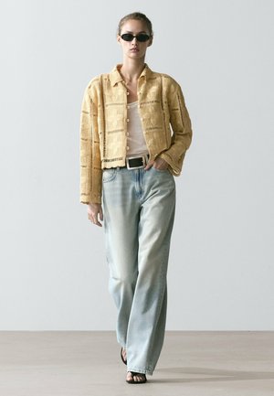 Donna con occhiali da sole neri, cardigan beige aperto a bottoni a maglia larga sopra una maglietta bianca, jeans a gamba larga azzurri, sandali neri e cintura nera.