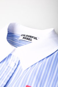 Blauw-wit gestreepte shirt met een klassieke kraag, met een stoffen label gemarkeerd "COLOURFUL REBEL" en een rode patch-detail.