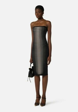 Donna che indossa un vestito midi a righe bianche e nere con sottili bretelle a catena dorata, che tiene una borsa nera e indossa tacchi alti neri.