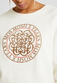 Primer plano de una persona que lleva un sudadera blanca con un diseño ornamental marrón circular y el texto "MOS MOSH X AÑOS" alrededor.