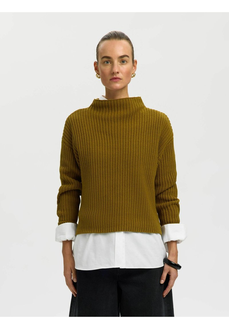 Femme portant un pull côtelé moutarde par-dessus une chemise blanche avec un pantalon noir, des boucles d'oreilles créoles en or et un bracelet noir, debout devant un fond blanc.