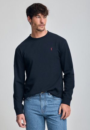 Zwarte lange mouwen t-shirt van een zachte katoenmix, met een rood geborduurd logo op de borst, gecombineerd met lichtblauwe spijkerbroek.