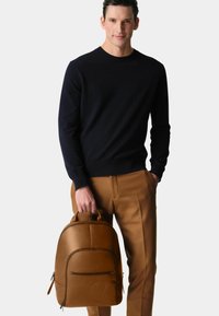 Brown leather backpack with rounded shape, front zip pocket, and embossed logo. Held by a person wearing a navy sweater and tan trousers.